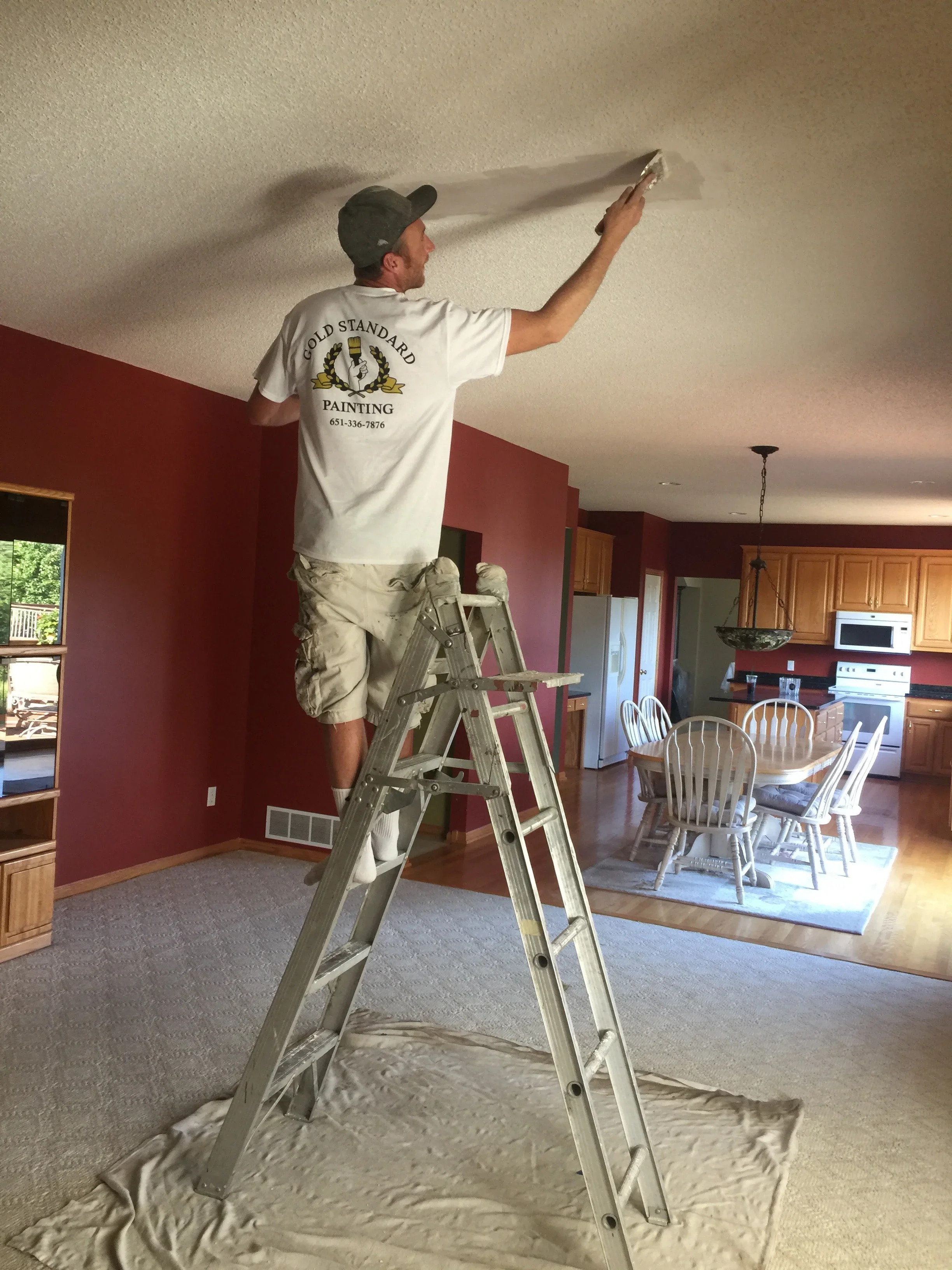Painter on ladder applying finish to ceiling with burgundy walls and wood cabinetry visible