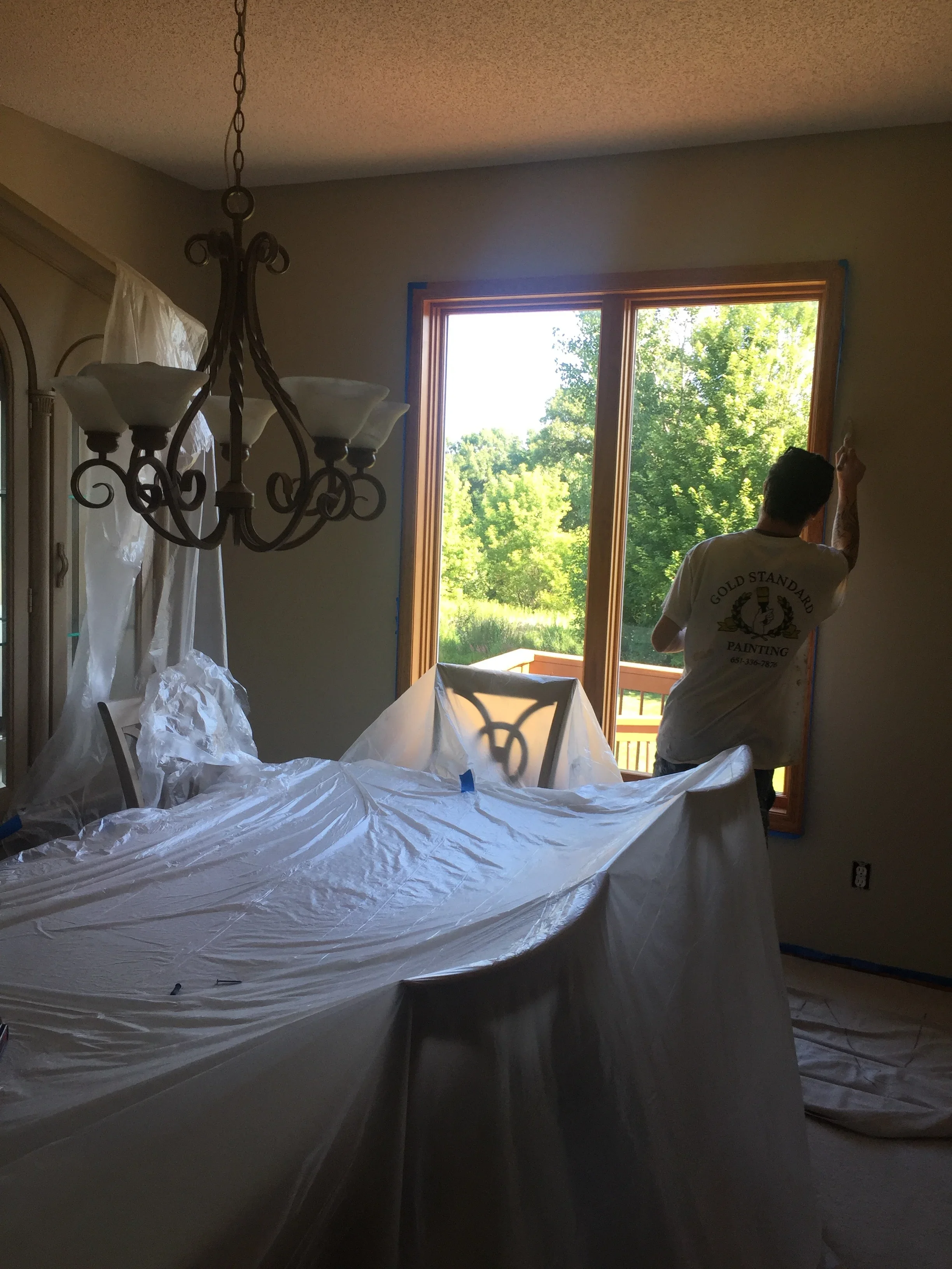 Interior room with plastic protection covering furniture and floors, painter working near large French doors with wood trim