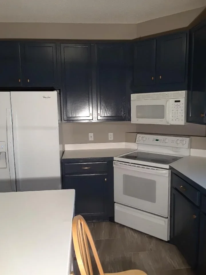 Navy blue painted kitchen cabinets with brass hardware, white appliances, and neutral countertops