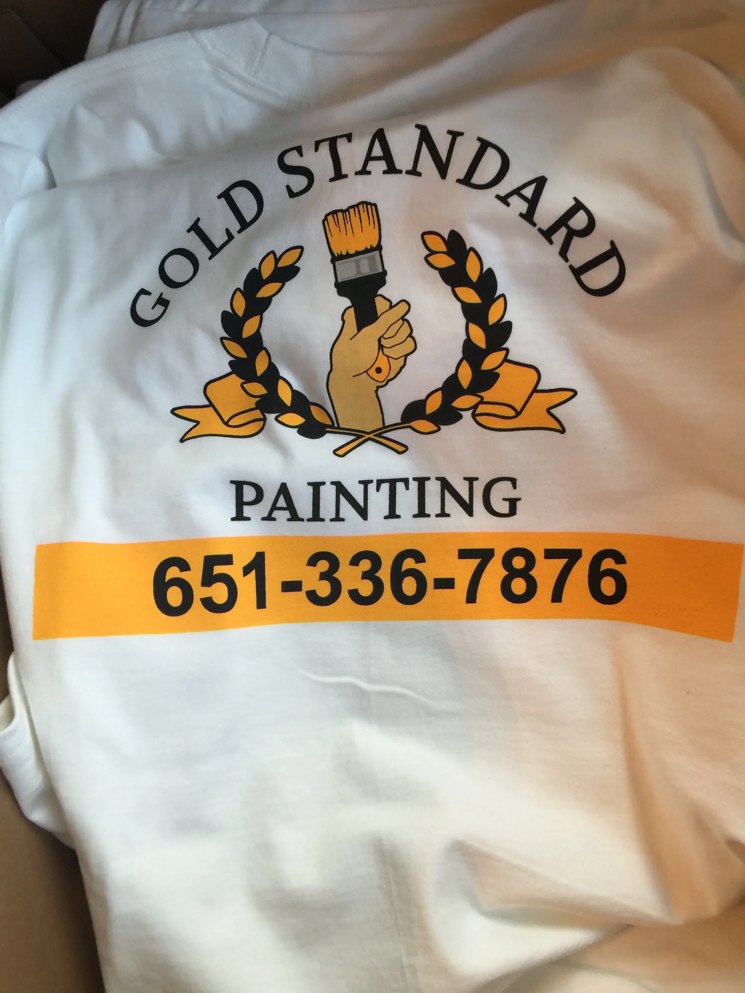 White t-shirt with gold and black painted logo design featuring paintbrush and laurel wreath motif