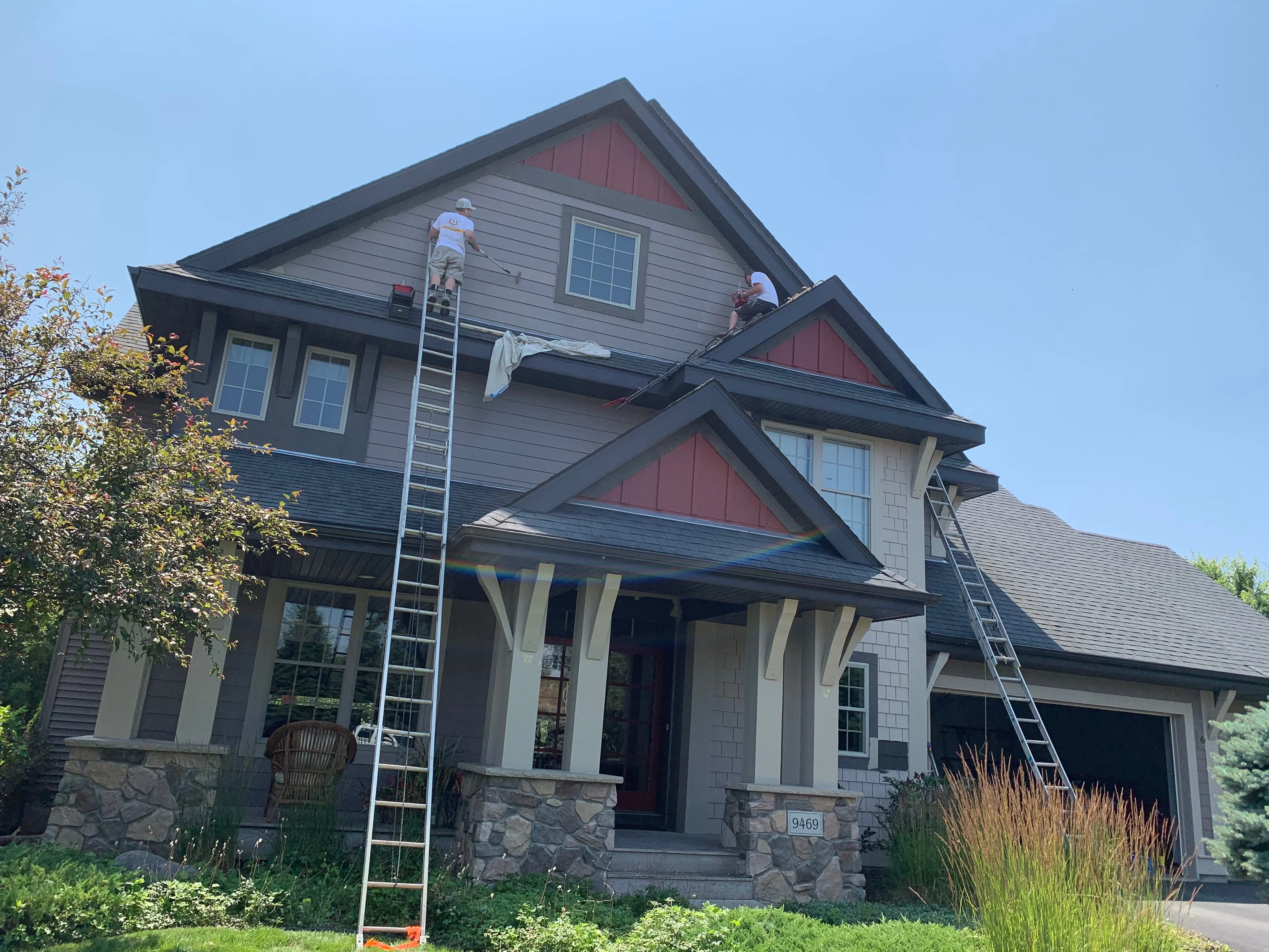 Two-story home with gray siding, burgundy gables, black trim, and stone foundation during exterior painting