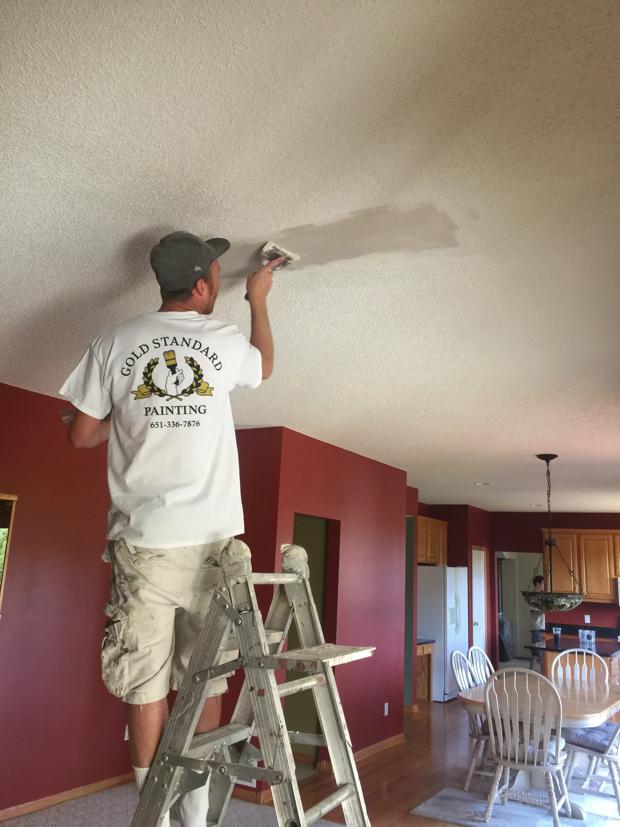 Painter on ladder finishing drywall ceiling seams with putty knife in residential interior