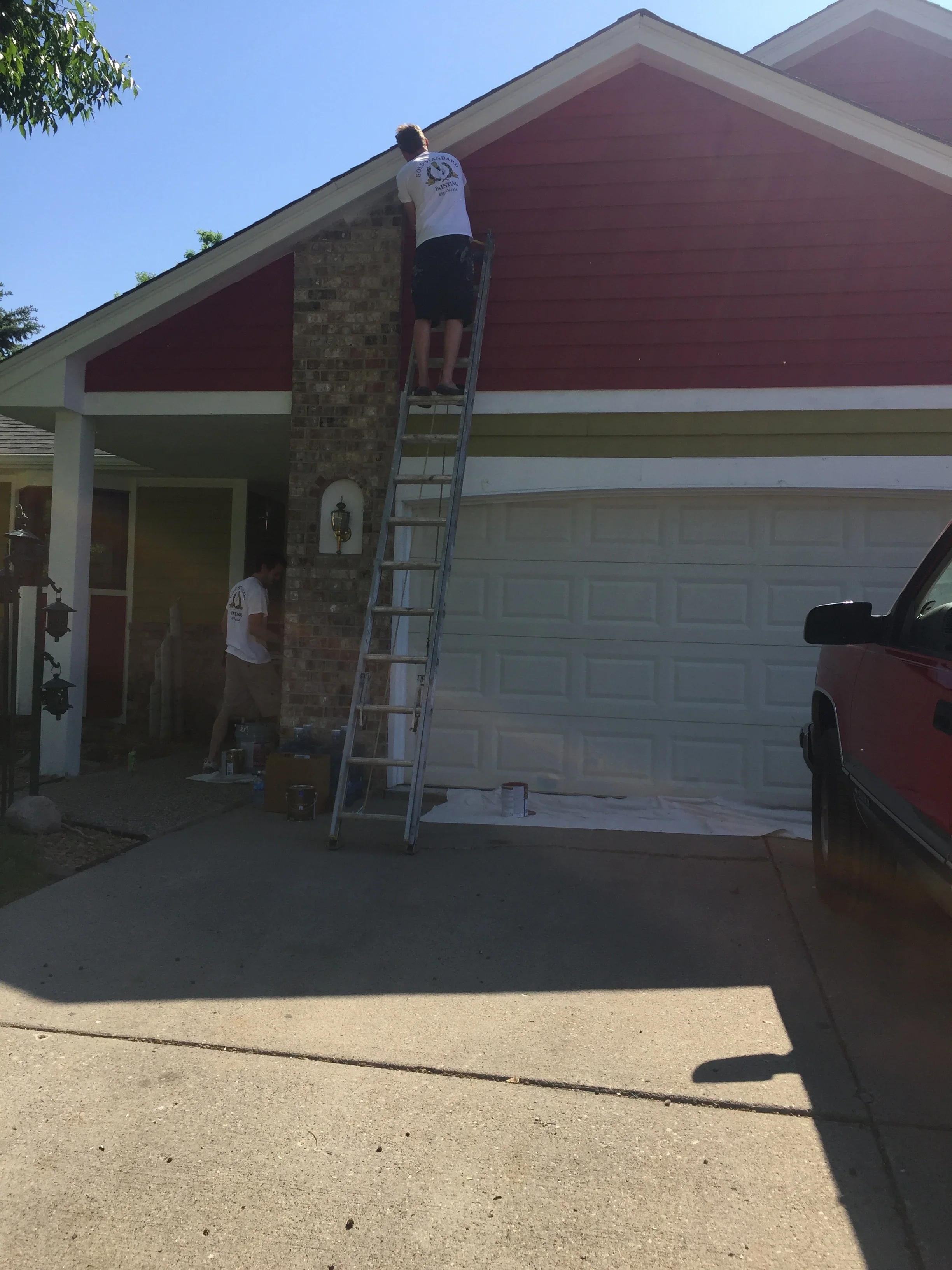 Painter on ladder applying burgundy paint to house exterior with white trim and brick chimney