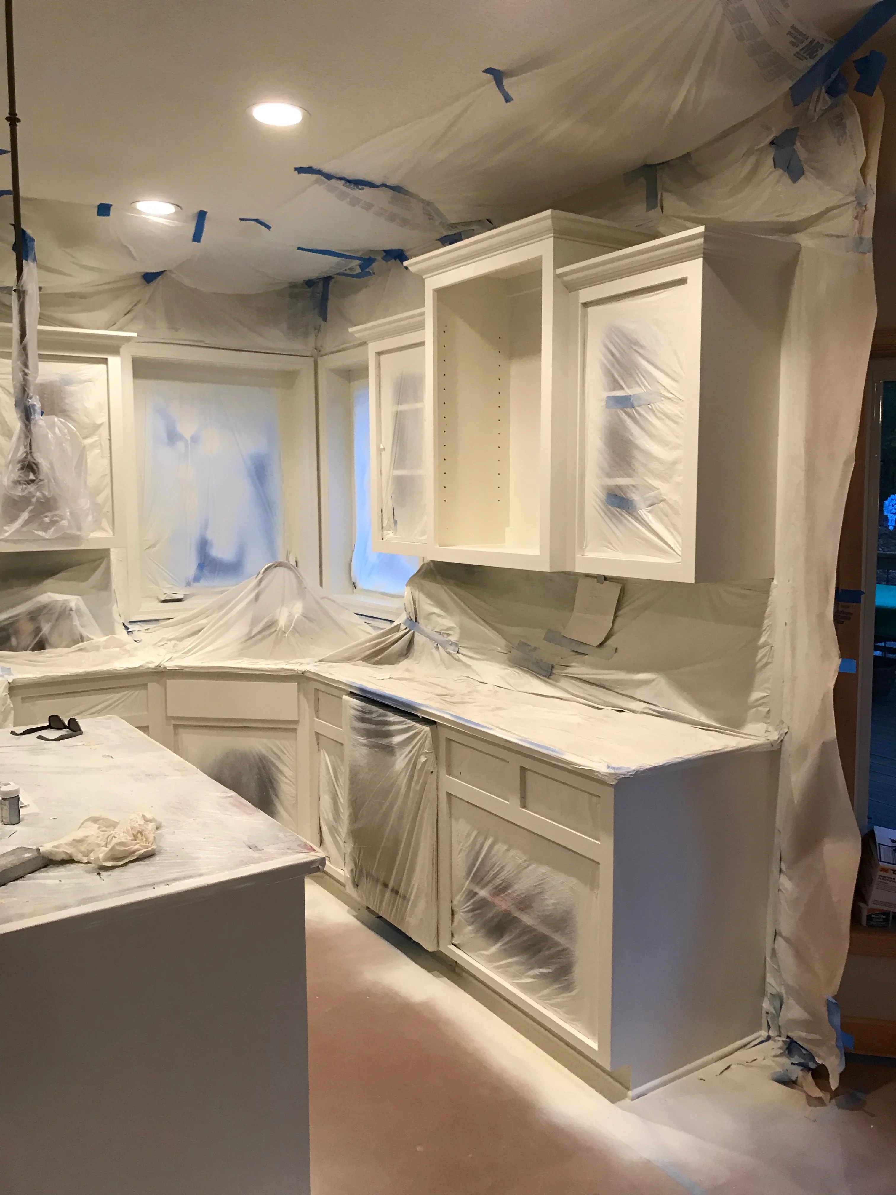 Kitchen cabinets masked with plastic sheeting and blue tape during painting preparation