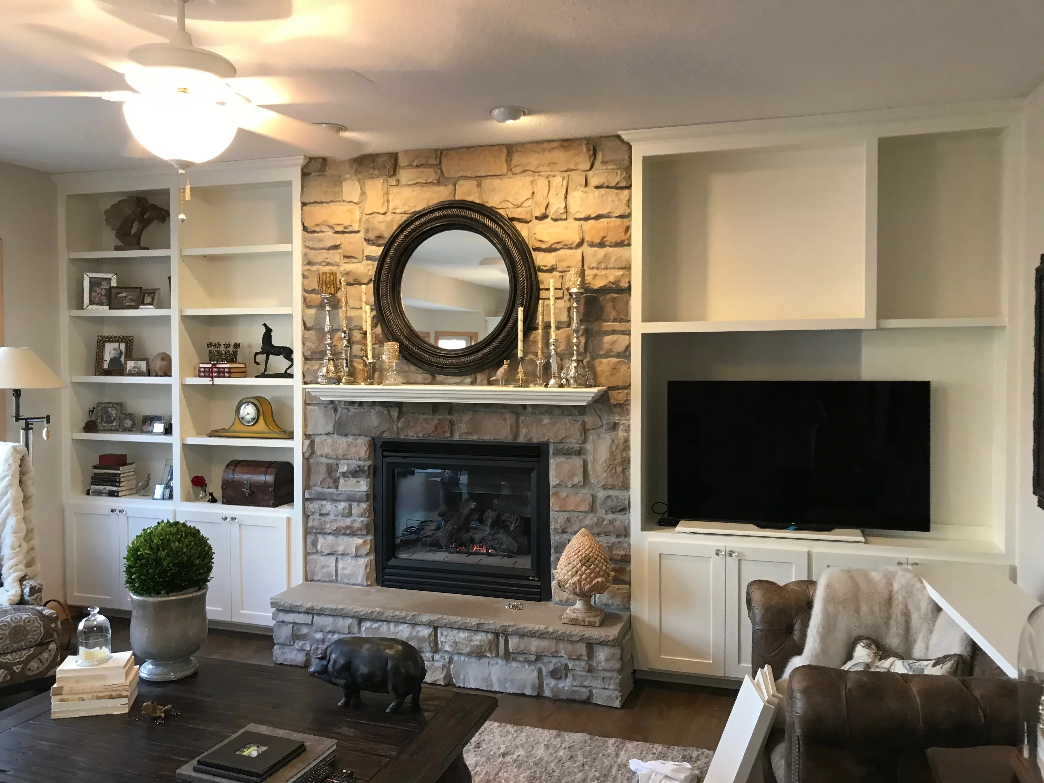 Stone fireplace with black gas insert, white built-in shelving, round mirror, and TV niche with painted cabinetry
