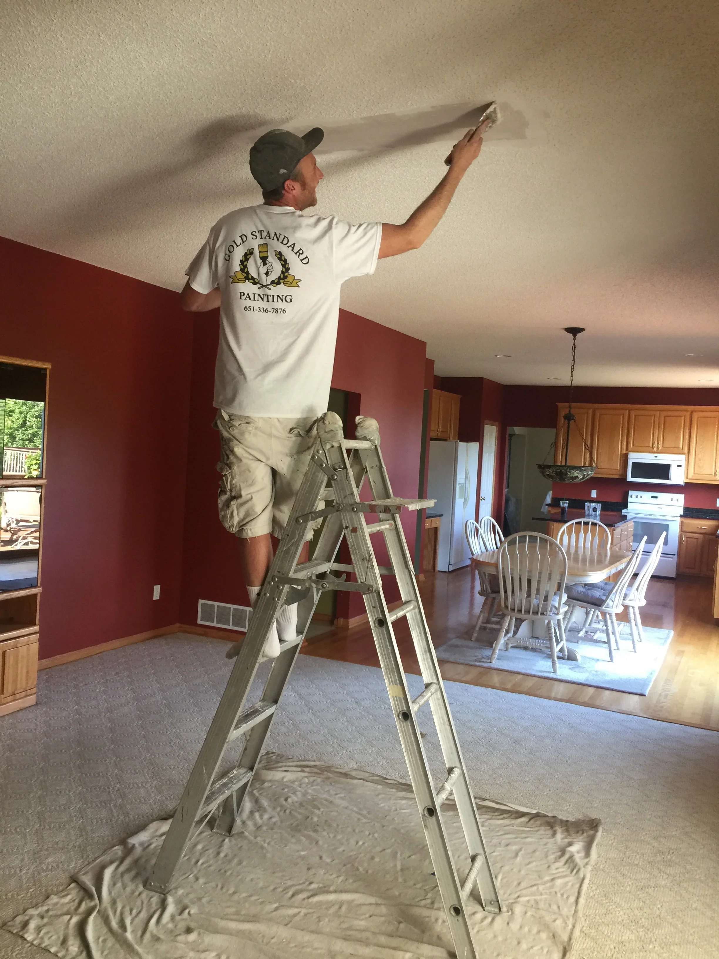Painter on ladder applying finish to ceiling with burgundy walls and wood cabinetry visible