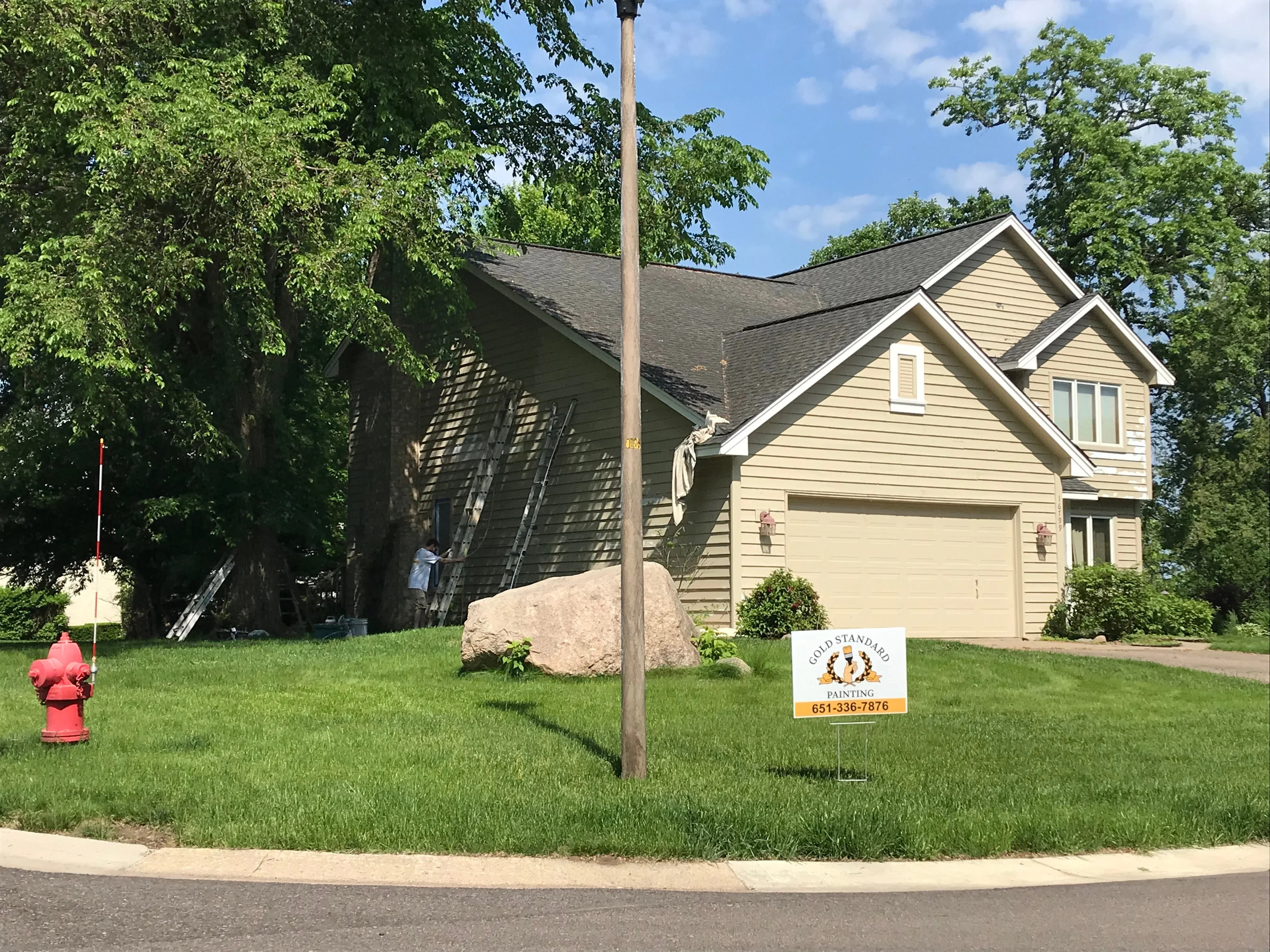 Two-story house with cream vinyl siding and white trim, exterior painting in progress with ladder setup